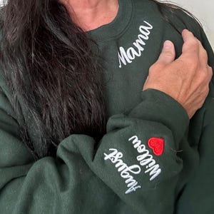 Custom embroidered Mama sweatshirt in forest green, featuring white stitched text on neckline and kids' names on the sleeve with a red heart. Personalized gift for moms, perfect for Mother's Day or birthdays. Cozy Gildan sweatshirt for proud mamas, showcasing children’s names in script font embroidery. Customizable and thoughtful apparel gift for mothers.