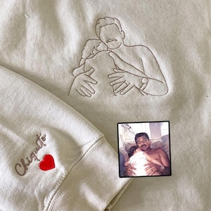 May include: A beige sweatshirt featuring brown thread embroidery of a person hugging a child. The sleeve displays the embroidered word Chiquito in brown thread next to a small, solid red heart. A small square photograph shows the original image.