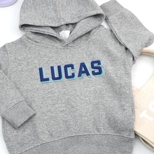 May include: A gray hooded sweatshirt with the name "LUCAS" embroidered in blue and light blue. The sweatshirt has a drawstring hood and long sleeves. The garment is made of a soft, comfortable material.
