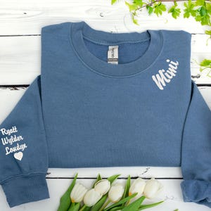 Custom embroidered sweatshirt in indigo blue featuring the word “Mimi” stitched near the neckline and grandkids’ names (Ryatt, Wylder, Londyn) with a heart on the sleeve. Personalized grandma gift with white thread on cozy Gildan crewneck. Perfect for Mother’s Day, birthday gifts for Mimi, or family keepsake sweatshirt. Laid flat with white tulips and spring greenery on a rustic white wooden background.