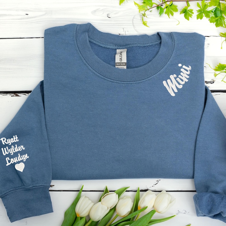 Custom embroidered sweatshirt in indigo blue featuring the word “Mimi” stitched near the neckline and grandkids’ names (Ryatt, Wylder, Londyn) with a heart on the sleeve. Personalized grandma gift with white thread on cozy Gildan crewneck. Perfect for Mother’s Day, birthday gifts for Mimi, or family keepsake sweatshirt. Laid flat with white tulips and spring greenery on a rustic white wooden background.