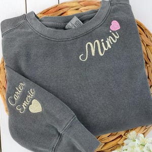 May include: A charcoal gray sweatshirt with embroidered text and heart accents. The word "Mimi" is embroidered on the chest, with a pink heart above it. The left sleeve features the names "Carter Emerie" and a gold heart.