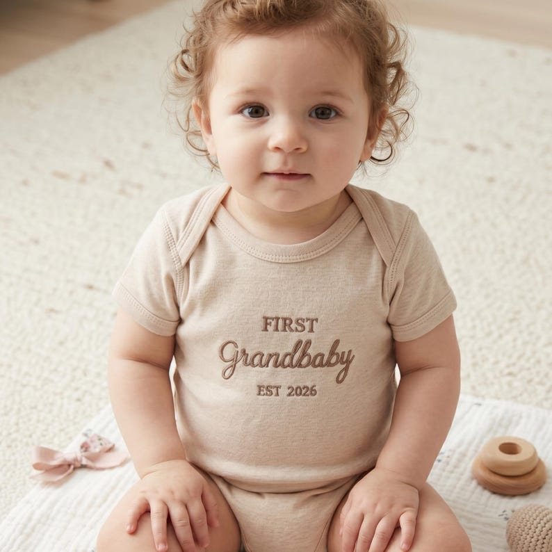 A cute baby sitting on a soft rug wearing a custom embroidered Natural bodysuit. The "First Grandbaby EST 2026" design is clearly visible, making it a heartwarming gift from grandparents. The Rabbit Skins 4400 model provides a comfortable, breathable fit, ideal for everyday wear and special family photo sessions in a bright, modern nursery.