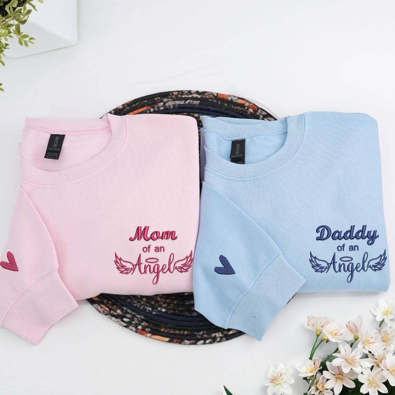 Flat lay of matching embroidered sweatshirts in light pink and light blue. The pink shirt reads “Mom of an Angel” and the blue shirt reads “Daddy of an Angel,” both with angel wing embroidery and small heart details on the sleeve. A touching memorial shirt set symbolizing everlasting love for a lost child.