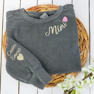 Pepper grey Comfort Colors sweatshirt with “Mimi” embroidered in cream script and a pink heart on the chest. Sleeve personalization features the names “Carter” and “Emerie” with a cream heart. Cozy and meaningful gift for grandmothers, Mother’s Day, or birthdays. High-quality custom embroidery makes this crewneck a cherished keepsake, perfect for Etsy shoppers seeking personalized family gifts.
