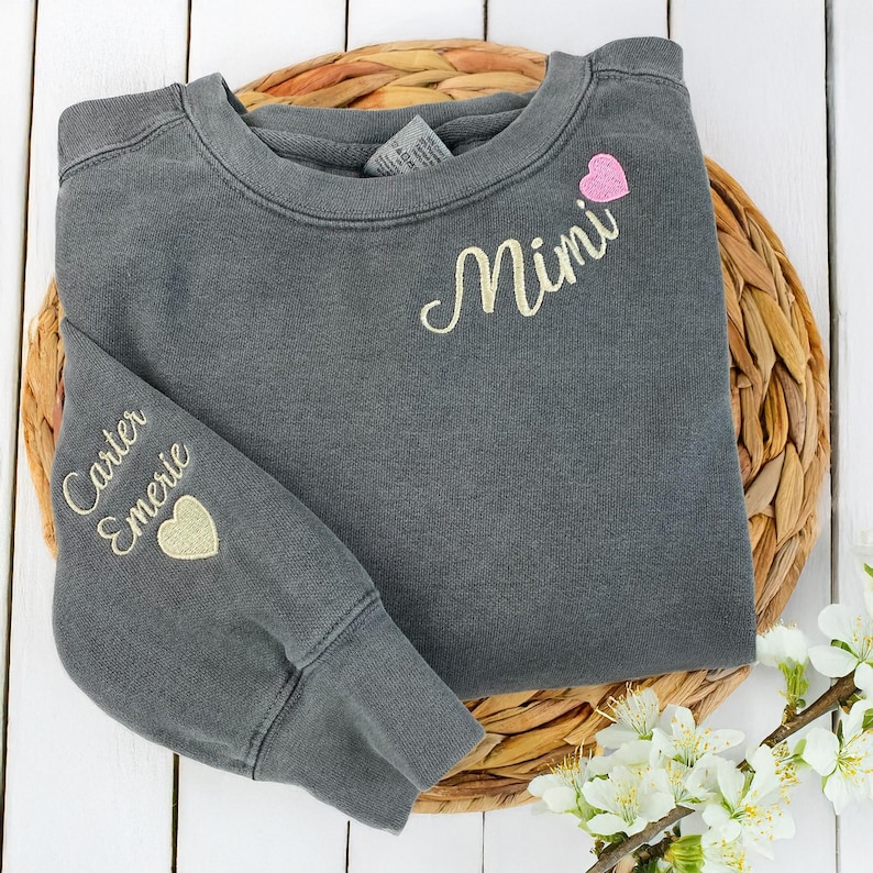 Pepper grey Comfort Colors sweatshirt with “Mimi” embroidered in cream script and a pink heart on the chest. Sleeve personalization features the names “Carter” and “Emerie” with a cream heart. Cozy and meaningful gift for grandmothers, Mother’s Day, or birthdays. High-quality custom embroidery makes this crewneck a cherished keepsake, perfect for Etsy shoppers seeking personalized family gifts.