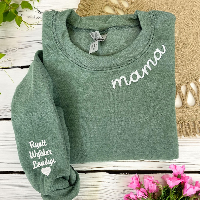 Custom embroidered heather dark green sweatshirt with 'mama' stitched on the neckline and children's names — Ryatt, Wylder, and Londyn — embroidered on the sleeve with a heart detail. Personalized Mother's Day gift, cozy mom sweater, perfect for mama bear gifts, mom of three sweatshirt, and custom name embroidery apparel. Flat lay style with floral accents, ideal for Etsy product listing.