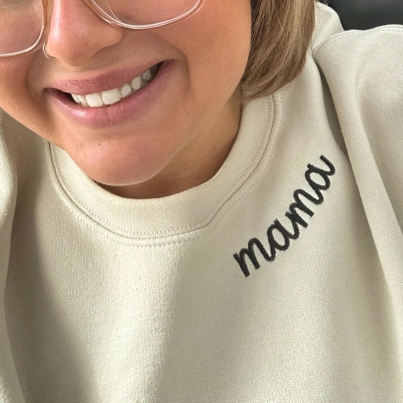 Close-up of a woman wearing a custom embroidered sand sweatshirt with the word "mama" stitched in black thread near the neckline. Cozy and minimalistic design perfect for personalized Mother’s Day gifts, new mom outfits, or everyday wear. Custom name placement adds sentimental touch. Great for Etsy shoppers seeking custom mama sweatshirts and embroidered gifts for moms.