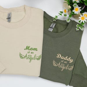 Set of sand and military green embroidered sweatshirts for mom and dad, featuring “Mom of an Angel” and “Daddy of an Angel” text in soft green and beige thread. Surrounded by white daisies, this cozy design conveys remembrance and love in an elegant, peaceful style.
