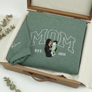 Könnte beinhalten: Ein grünes Sweatshirt mit dem Wort "MOM" in weißen Blockbuchstaben. Das Sweatshirt zeigt ein gesticktes Bild einer Mutter, die ein Baby hält, mit "EST. 2025" darunter. Der Ärmel hat den Namen "Kane" in Weiß gestickt.