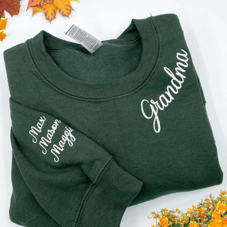 Forest green embroidered sweatshirt folded neatly, featuring "Grandma" stitched on the neckline and grandkids' names "Max, Mason, Maggie" on the sleeve with a white heart—perfect personalized gift for grandma. Custom embroidery design ideal for Mother’s Day, Christmas, or birthday. Displayed on white background with fall leaves and yellow-orange flowers for a cozy seasonal vibe.