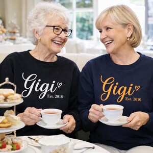 Op de afbeelding: Twee vrouwen dragen zwarte en marineblauwe sweatshirts met het woord "Gigi" in script en het oprichtingsjaar. Ze houden theekopjes vast en glimlachen. Het zwarte sweatshirt zegt "Est. 2018" en het marineblauwe sweatshirt zegt "Est. 2010".