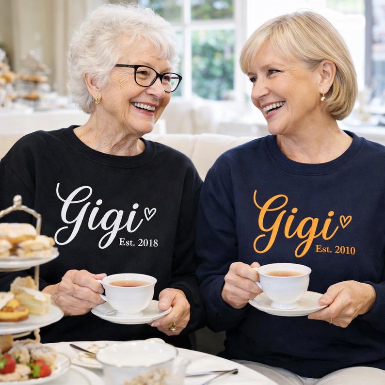 Op de afbeelding: Twee vrouwen dragen zwarte en marineblauwe sweatshirts met het woord "Gigi" in script en het oprichtingsjaar. Ze houden theekopjes vast en glimlachen. Het zwarte sweatshirt zegt "Est. 2018" en het marineblauwe sweatshirt zegt "Est. 2010".