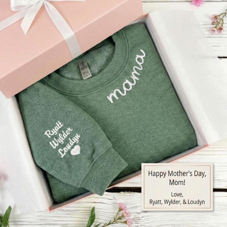 May include: A green sweatshirt with the word "mama" embroidered in white, nestled in a pink gift box tied with a white ribbon. The sleeve features embroidered names and a heart. A card reads "Happy Mother's Day, Mom!"