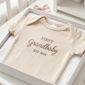 A personalized "First Grandbaby" baby bodysuit in Natural neatly folded inside a premium gift box. Accompanied by a small lamb toy and baby books, this Rabbit Skins 4400 onesie makes for a luxury baby shower gift set. The minimalist design and custom embroidery cater to the growing trend of aesthetic and thoughtful newborn presents.