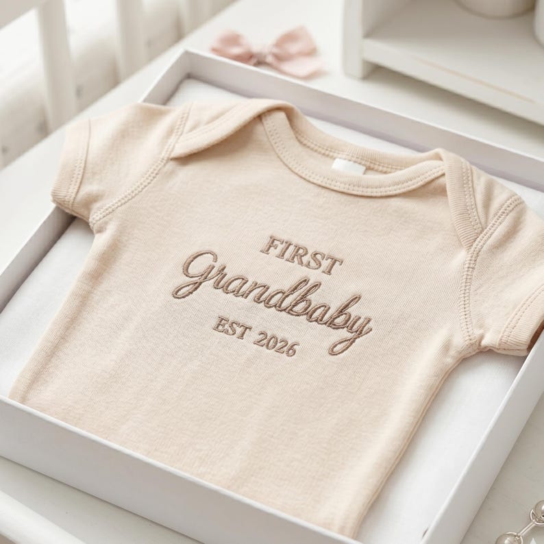 A personalized "First Grandbaby" baby bodysuit in Natural neatly folded inside a premium gift box. Accompanied by a small lamb toy and baby books, this Rabbit Skins 4400 onesie makes for a luxury baby shower gift set. The minimalist design and custom embroidery cater to the growing trend of aesthetic and thoughtful newborn presents.