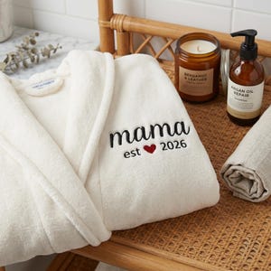 May include: A white terry cloth robe with the word "mama" embroidered in black script, a red heart, and the year "2026". A candle, shampoo bottle, and rolled towel are on a wicker tray.