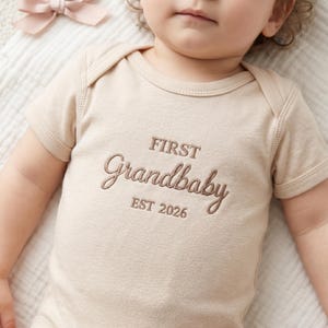 An infant lying down wearing a personalized "First Grandbaby" bodysuit in Natural. Surrounded by aesthetic baby accessories like wooden rattles, leather shoes, and dried flowers. This Rabbit Skins 4400 onesie is perfect for newborn photography and milestone announcements, offering a cozy and stylish fit for the new family addition.