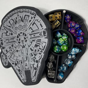 May include: A black and silver dice tray shaped like the Millennium Falcon from Star Wars. The tray has seven compartments for dice, with one compartment featuring a silver metal dice tower. The tray is open and shows a variety of colorful dice.
