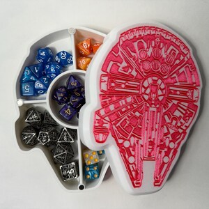 May include: A white dice tray shaped like the Millennium Falcon from Star Wars. The tray has six compartments for different types of dice. The compartments are filled with various colored dice, including blue, orange, and silver.