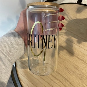 May include: A clear glass tumbler with a bamboo lid and a black and gold sticker that says "BRITNEY". A clear drinking straw is in the tumbler.