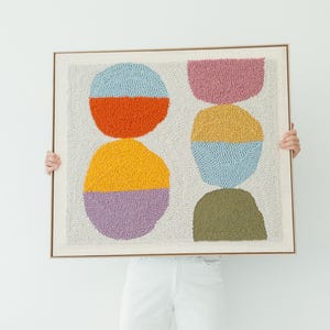 Può includere: Un'opera d'arte tessile astratta incorniciata con forme arrotondate colorate in blu, arancione, giallo, viola, rosa e verde. L'opera d'arte è tenuta su uno sfondo bianco. Un vaso marrone con fiori secchi si trova nell'angolo in basso a sinistra.