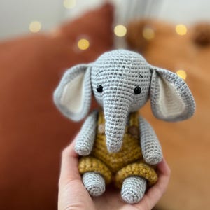 May include: A grey crocheted elephant toy wearing a yellow crocheted jumpsuit. The elephant has black eyes and a small, grey trunk.
