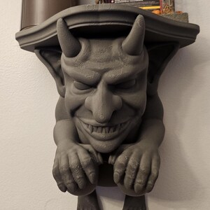 May include: A gray, devil-themed shelf bracket with a grinning face, horns, and clawed hands and feet. The shelf has a curved design and is mounted on a wall. The bracket is made of a solid material.