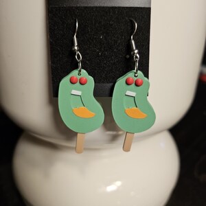 May include: A pair of green and orange popsicle earrings with red eyes and a white smile. The earrings are on a black card with a hole in the center.