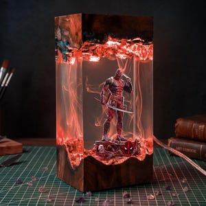 May include: A rectangular resin and wood lamp featuring a Deadpool figurine standing on a base with the character logo. The clear resin section is illuminated with red light, creating a smoke-like effect around the figure holding a sword.