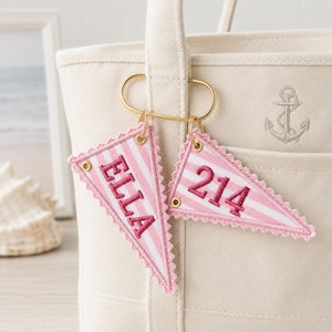 Peut inclure: Un sac fourre-tout en toile couleur crème avec un logo ancre brodé et deux porte-clés en forme de fanion triangulaire fixés à un anneau en métal doré. Les fanions sont rayés rose clair et blanc avec le texte ELLA et 214 en rose foncé.