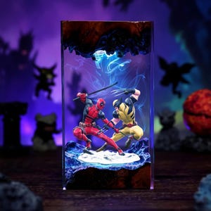 May include: A rectangular resin block containing a miniature diorama of Deadpool and Wolverine in a combat pose. Deadpool wears a red and black suit with a katana, while Wolverine wears a yellow and black suit with extended metal claws.