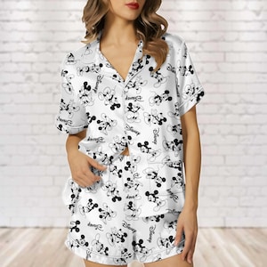 Könnte beinhalten: Weißes Satin-Pyjama-Set mit schwarzem Mickey Mouse-Muster. Das Set besteht aus einem kurzärmeligen Hemd mit Knöpfen und passenden Shorts. Das Wort "Disney" ist ebenfalls auf dem Pyjama aufgedruckt.