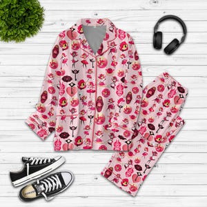 May include: Pink pyjama set featuring a repeating pattern of cartoon characters, keys, and hearts. The long-sleeved top has a notched collar and button-down front. The trousers have an elasticated waistband. Black Converse trainers are at the bottom.