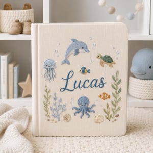 Könnte beinhalten: Ein cremefarbenes, stoffbezogenes Erinnerungsbuch mit gestickten Meeresbewohnern wie einem Delfin, einer Qualle, einem Oktopus, einer Meeresschildkröte und einem Fisch. Der Name Lucas ist mittig in blauer Schreibschrift gestickt, umgeben von Algen.