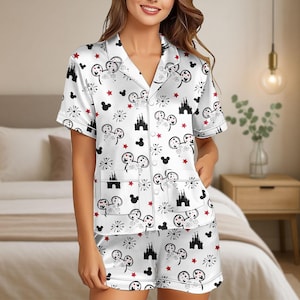 Könnte beinhalten: Weißes Satin-Pyjama-Set mit einem Muster aus Mickey Mouse-Köpfen, Feuerwerk, roten Sternen und Schlosssilhouetten. Das kurzärmelige Oberteil hat einen Reverskragen und Vordertaschen. Die Shorts haben einen elastischen Bund.