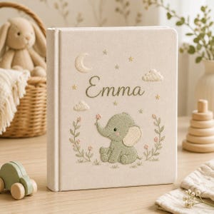 Könnte beinhalten: Ein cremefarbenes, stoffbezogenes Buch mit Stickereien, die eine Mondsichel, Sterne, Wolken und einen grünen Elefanten zeigen. Der Name Emma ist in Schreibschrift über dem Elefanten eingestickt, der von zwei Blumenranken umrahmt wird.