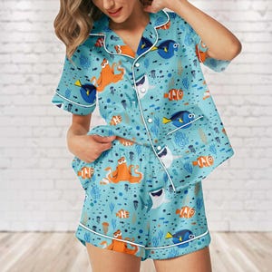 Puede incluir: Conjunto de pijama azul claro con un estampado de vida marina. La parte superior de manga corta y los pantalones cortos presentan peces de dibujos animados, pulpos y tiburones. Ribetes blancos delinean los bordes del pijama.