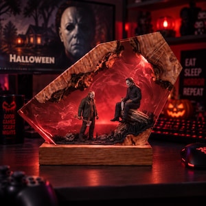 May include: A decorative lamp featuring a live-edge wood frame filled with red resin, housing miniature figures of Jason Voorhees holding an axe and Michael Myers holding a knife. The piece is mounted on a rectangular wooden base with internal lighting.