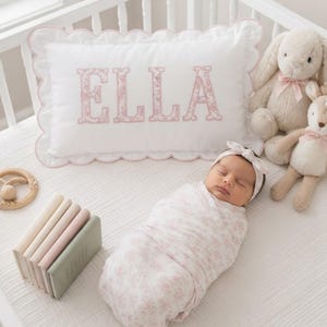 May include: A white rectangular pillow with a scalloped edge featuring the name ELLA in pink floral letters. A swaddled infant wears a matching pink floral headband. Nearby are two plush animal toys, a wooden ring, and a stack of four small books.