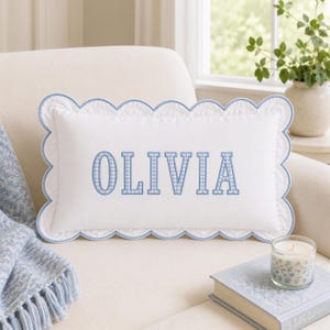 Könnte beinhalten: Ein rechteckiges weißes Zierkissen mit gewelltem Rand, der mit hellblauer Paspelierung versehen ist. In der Mitte des Kissens ist der Name OLIVIA in einem hellblau-weiß karierten Vichy-Muster in einer fetten Blockschrift eingestickt.