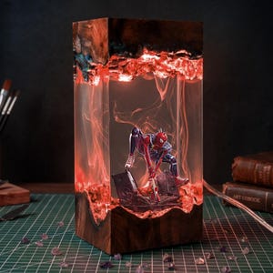 Könnte beinhalten: Eine rechteckige Lampe aus dunklem Holz und klarem Harz mit einem beleuchteten roten Innenraum. Im Harz befindet sich eine Miniaturfigur eines Superhelden im rot-blauen Anzug, die auf einem Sockel mit zwei kleinen Lautsprechern und einer Gitarre hockt.