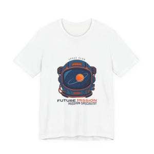 May include: White t-shirt featuring a graphic of an astronaut helmet with a planet and rings inside the visor. The design includes the text "SPACE CLUB" above the helmet and "FUTURE MISSION MISSION SPECIALIST" below.
