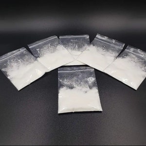 May include: Seven clear plastic bags filled with a white powder substance. The bags are arranged in a circle on a black surface.