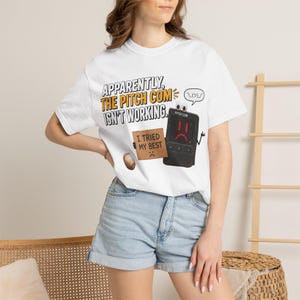 Op de afbeelding: Een wit t-shirt met korte mouwen en een afbeelding van een zwart elektronisch apparaat met een verdrietig gezicht, dat een bruin bordje vasthoudt met de tekst I TRIED MY BEST. Erboven staat APPARENTLY, THE PITCH COM ISN'T WORKING met een emoticon.