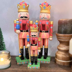 May include: Three wooden nutcrackers in a row, each with a crown and mustache. The nutcrackers are painted in shades of red and pink with gold accents. They stand on green bases and hold staffs. A festive holiday decoration.