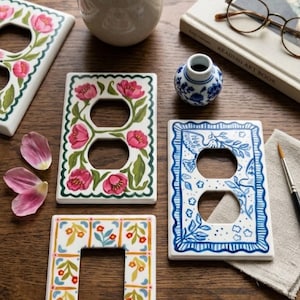 Puede incluir: Una colección de cubiertas decorativas de cerámica para enchufes con diseños florales y botánicos. Las cubiertas presentan tulipanes rosas pintados a mano, pájaros azules y otros patrones coloridos. También hay un pequeño jarrón con flores, un libro y un pincel.