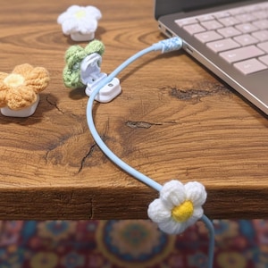 May include: A light blue charging cable connected to a laptop, featuring a white daisy-shaped crochet cable tidy. Other crochet flower cable clips in white, yellow, and green are also visible.