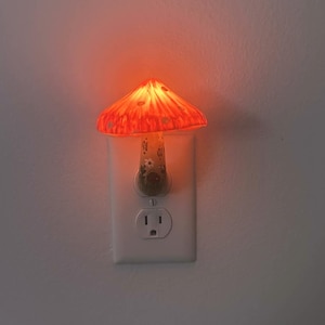 Peut inclure: Veilleuse décorative en forme de champignon avec une lueur orange chaude. Le chapeau du champignon présente un motif texturé et la base est conçue pour être branchée dans une prise murale standard. La veilleuse est fixée sur un mur blanc.