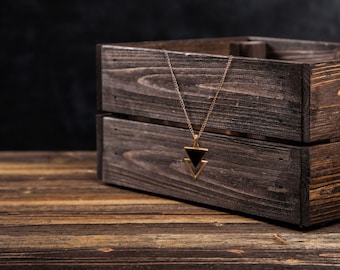 Puede incluir: Un collar dorado con un colgante triangular negro cuelga frente a una caja de madera rústica. El collar tiene una cadena delicada y un diseño geométrico. La caja está hecha de madera marrón oscura y se encuentra sobre una superficie de madera.